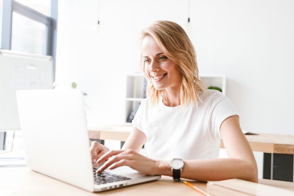 woman-smiling-whilst-sitting-down-to-take-online-exam-on-her-laptop